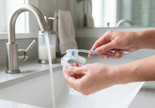 A close-up of hands cleaning a clear night guard with a toothbrush under running water in a bathroom.