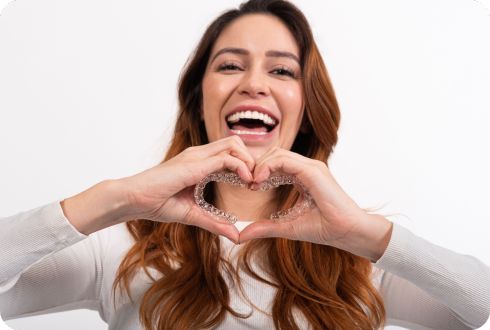 A woman smiling while holding clear aligners in both hands to create a heart shape